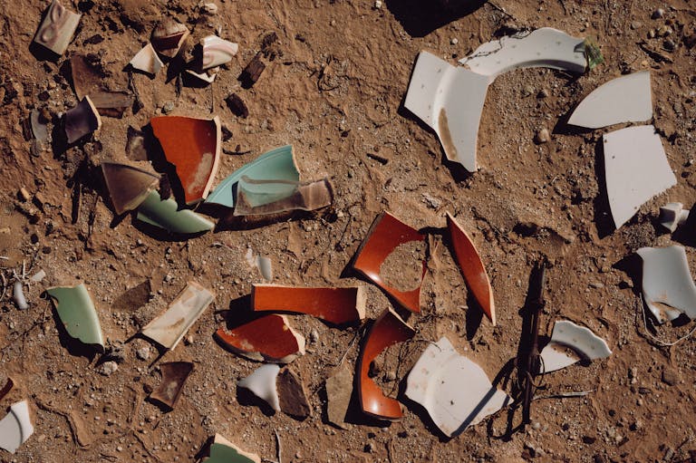 A collection of broken ceramic pieces scattered on a dirt ground under sunlight.